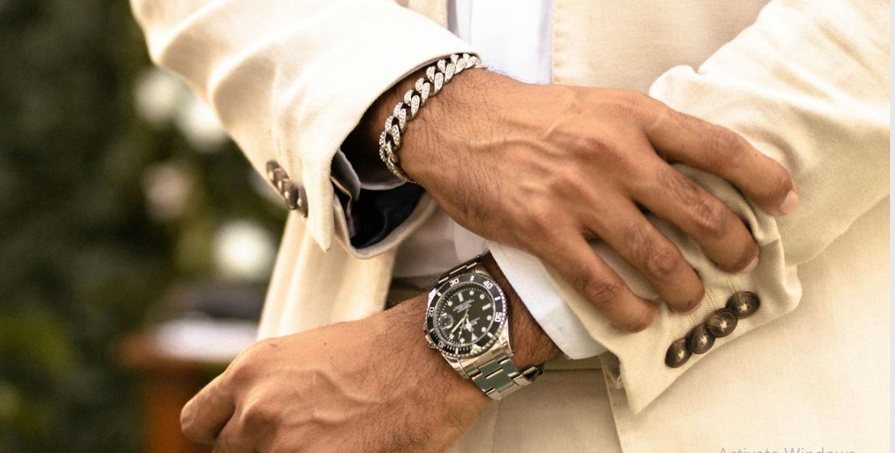 Un hombre vistiendo un elegante traje blanco crema y mostrando un reloj de pulsera y una pulsera con estilo en su mano.